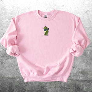 Puede incluir: Una sudadera rosa claro con cuello redondo y mangas largas. La sudadera presenta un peque&ntilde;o bordado de una rana con una bufanda azul. La rana es verde y blanca con detalles negros. La sudadera est&aacute; hecha de un material suave.