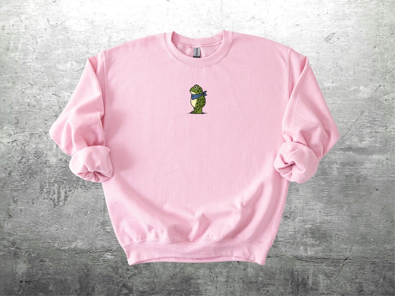 Puede incluir: Una sudadera rosa claro con cuello redondo y mangas largas. La sudadera presenta un peque&ntilde;o bordado de una rana con una bufanda azul. La rana es verde y blanca con detalles negros. La sudadera est&aacute; hecha de un material suave.