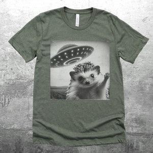 May include: A green t-shirt with a black and white image of a hedgehog waving at a flying saucer.