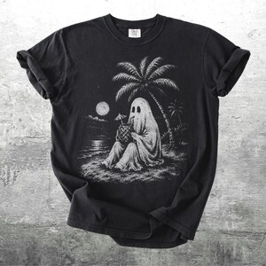 May include: A black short-sleeve t-shirt featuring a white graphic illustration of a ghost sitting on a beach under a palm tree at night. The ghost is holding a pineapple drink with a small umbrella. A full moon is visible in the sky above the water.