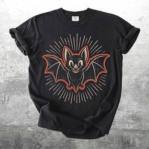 May include: A black short-sleeve t-shirt featuring a centered graphic of a smiling bat with orange outlines and wings spread wide. The bat is surrounded by a sunburst pattern of thin white lines. The design has a distressed, vintage-inspired texture.