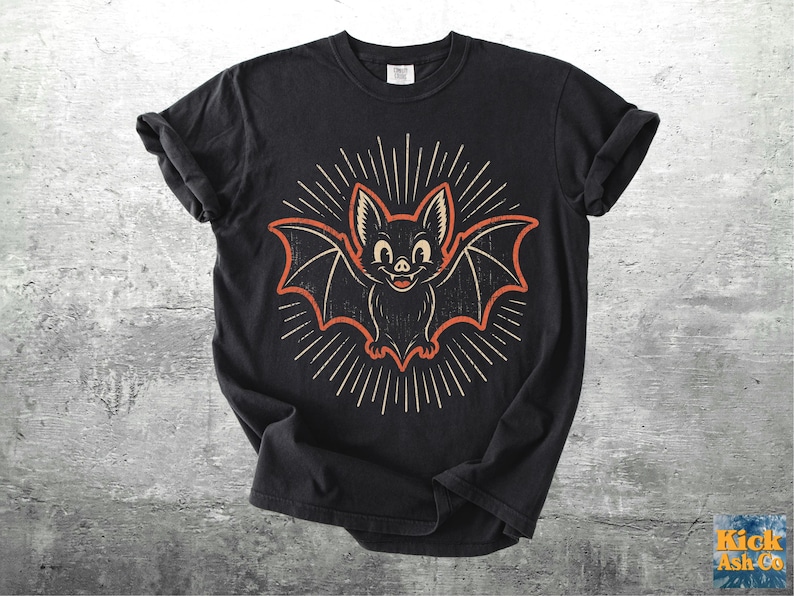 May include: A black short-sleeve t-shirt featuring a centered graphic of a smiling bat with orange outlines and wings spread wide. The bat is surrounded by a sunburst pattern of thin white lines. The design has a distressed, vintage-inspired texture.