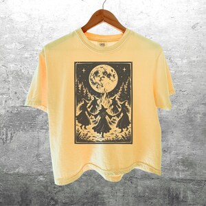 May include: A pale yellow short-sleeve t-shirt featuring a dark brown graphic print of four figures in pointed hats dancing in a circle around a campfire under a large full moon, surrounded by pine trees and stars, hanging on a wooden hanger.