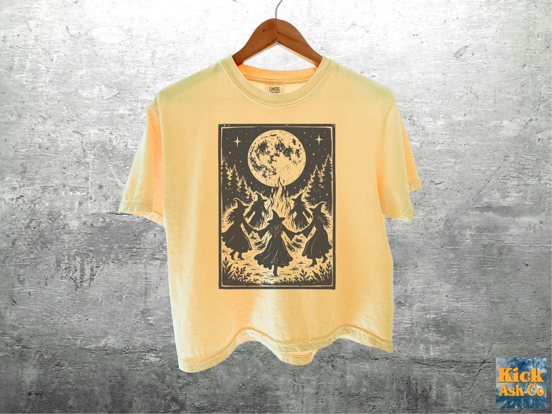 May include: A pale yellow short-sleeve t-shirt featuring a dark brown graphic print of four figures in pointed hats dancing in a circle around a campfire under a large full moon, surrounded by pine trees and stars, hanging on a wooden hanger.
