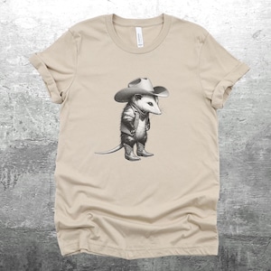 May include: A beige t-shirt with a graphic of a possum wearing a cowboy hat and boots. The possum is looking to the left.