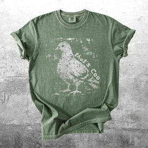 May include: Olive green t-shirt with a distressed white graphic of a pigeon and the text "that's Coo". The shirt has short sleeves and a crew neck. The fabric appears soft and slightly faded.