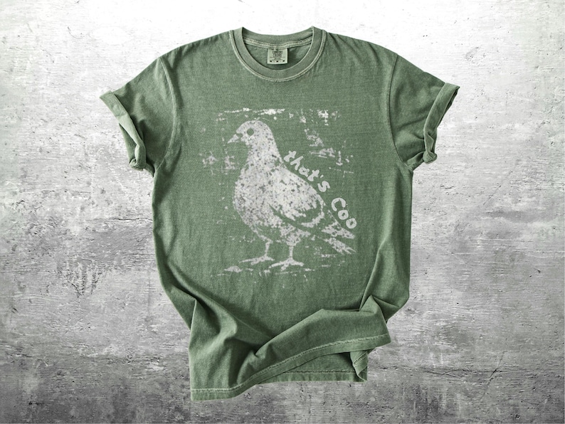 May include: Olive green t-shirt with a distressed white graphic of a pigeon and the text "that's Coo". The shirt has short sleeves and a crew neck. The fabric appears soft and slightly faded.