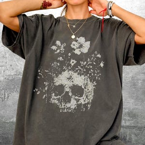 Pu&ograve; includere: T-shirt grigio scuro con un design a teschio e fiori bianchi. La stampa grafica bianca contrasta con il tessuto grigio. La maglietta ha maniche corte e una vestibilit&agrave; ampia, ideale per un abbigliamento casual.