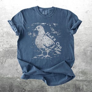 May include: A blue t-shirt with a distressed white graphic of a pigeon and the text "that's Coo". The shirt has rolled-up sleeves and a crew neck. The fabric appears to be soft and pre-shrunk.