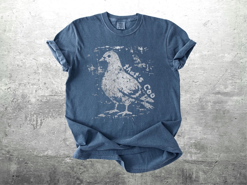 May include: A blue t-shirt with a distressed white graphic of a pigeon and the text "that's Coo". The shirt has rolled-up sleeves and a crew neck. The fabric appears to be soft and pre-shrunk.
