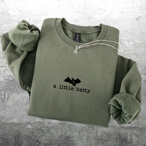 May include: Olive green sweatshirt with the phrase "a little batty" and a bat graphic. A silver necklace with a small charm is draped across the front. The sweatshirt has long sleeves and a crew neck.