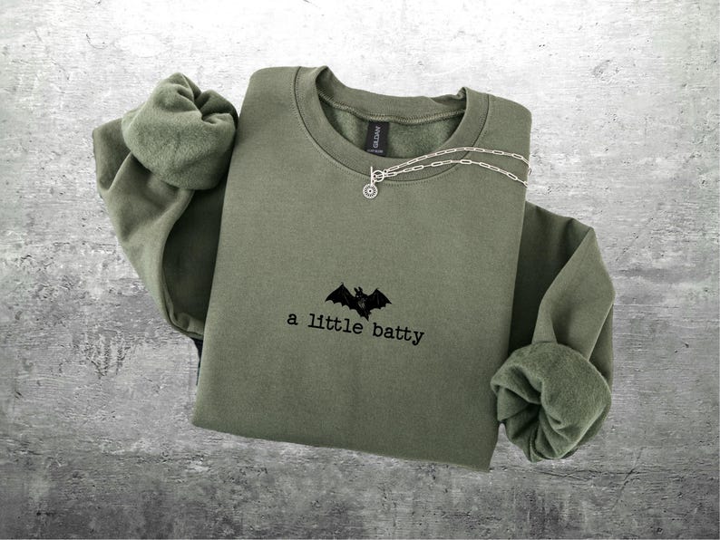 May include: Olive green sweatshirt with the phrase "a little batty" and a bat graphic. A silver necklace with a small charm is draped across the front. The sweatshirt has long sleeves and a crew neck.