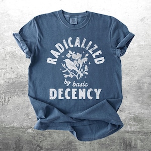 May include: A blue t-shirt with rolled-up sleeves. The shirt features the white text "RADICALIZED" above a bird and floral design, with "by basic DECENCY" below. The shirt is made of a soft material.
