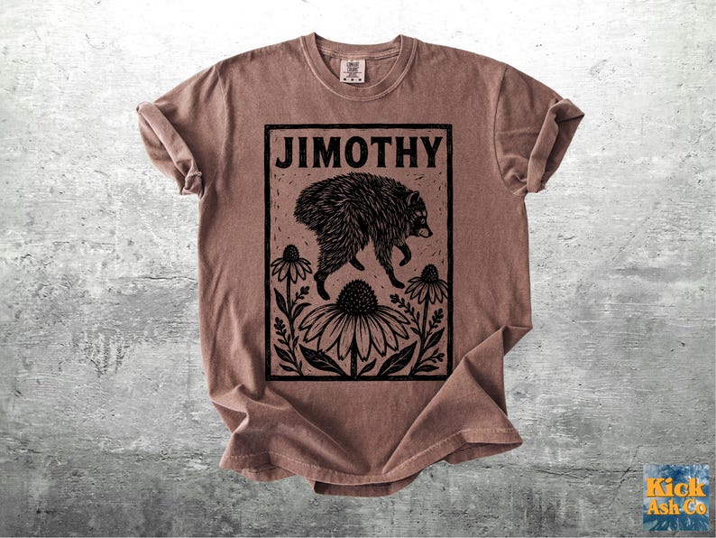 Peut inclure: Un t-shirt couleur vieux rose avec une impression graphique noire style encre. Le motif repr&eacute;sente un raton laveur sautant au-dessus de trois grandes fleurs, encadr&eacute; par un rectangle avec le texte JIMOTHY en lettres capitales grasses.