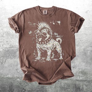 May include: Brown t-shirt featuring a distressed graphic of a punk rock pug dog with a mohawk and studded collar. The dog is in a standing pose, with its tongue sticking out. The shirt has rolled-up sleeves.