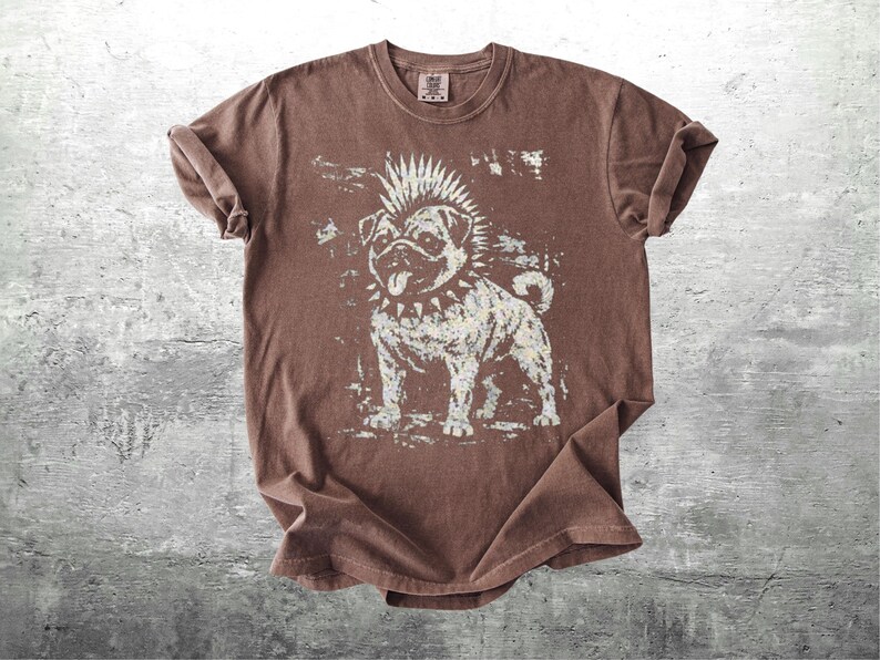May include: Brown t-shirt featuring a distressed graphic of a punk rock pug dog with a mohawk and studded collar. The dog is in a standing pose, with its tongue sticking out. The shirt has rolled-up sleeves.
