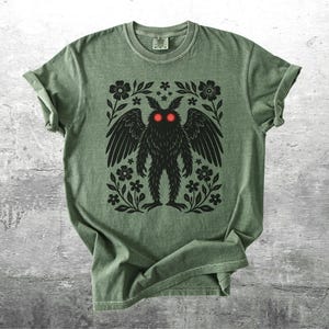 May include: Sage green t-shirt featuring a black illustration of a winged creature with red eyes, surrounded by floral and leaf accents. The design is centered on the shirt, creating a striking visual.
