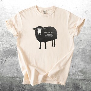 Puede incluir: Camiseta color crema con un gr&aacute;fico de oveja negra y el texto "EMBRACE WHAT MAKES YOU UNIQUE". La ilustraci&oacute;n de la oveja est&aacute; centrada en la camiseta. La camiseta tiene mangas cortas y cuello redondo.
