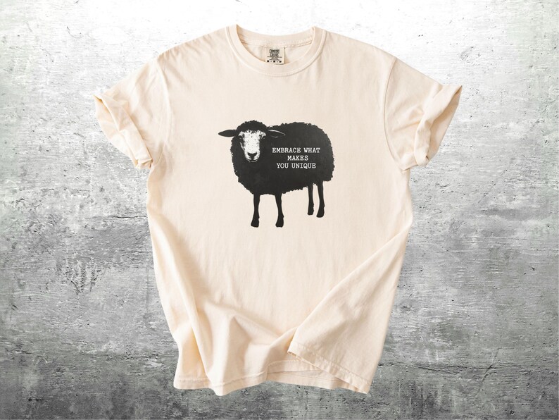 Puede incluir: Camiseta color crema con un gr&aacute;fico de oveja negra y el texto "EMBRACE WHAT MAKES YOU UNIQUE". La ilustraci&oacute;n de la oveja est&aacute; centrada en la camiseta. La camiseta tiene mangas cortas y cuello redondo.