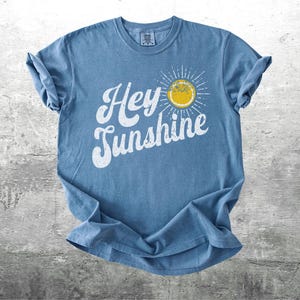 May include: A blue t-shirt with the words "Hey Sunshine" in white script and a yellow sun graphic. The shirt is folded and laid on a gray surface. The shirt has a crew neck and short sleeves.