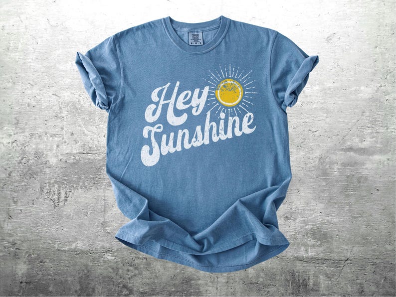 May include: A blue t-shirt with the words "Hey Sunshine" in white script and a yellow sun graphic. The shirt is folded and laid on a gray surface. The shirt has a crew neck and short sleeves.