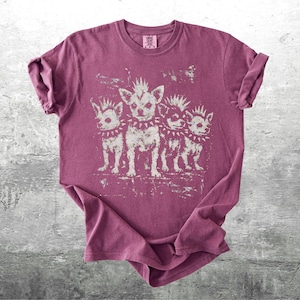 May include: A maroon t-shirt featuring a distressed graphic of four chihuahua dogs with mohawks and studded collars. The dogs are standing in a row. The shirt is folded on a gray surface.