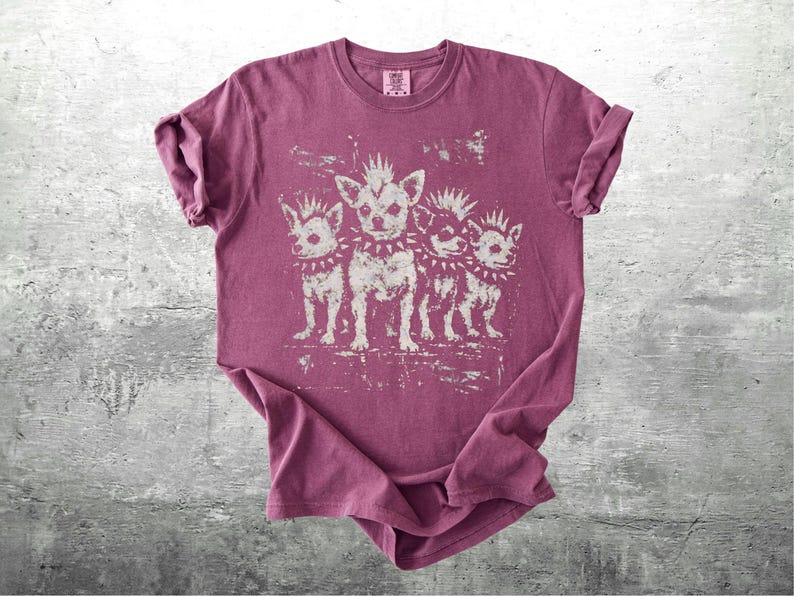 May include: A maroon t-shirt featuring a distressed graphic of four chihuahua dogs with mohawks and studded collars. The dogs are standing in a row. The shirt is folded on a gray surface.