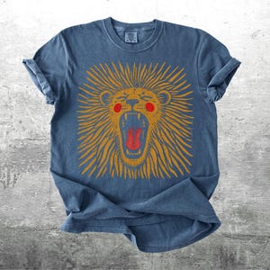 May include: A blue t-shirt featuring a distressed lion design. The lion has an open mouth, orange mane, red tongue, and red cheeks. The shirt has rolled-up sleeves and a crew neck.