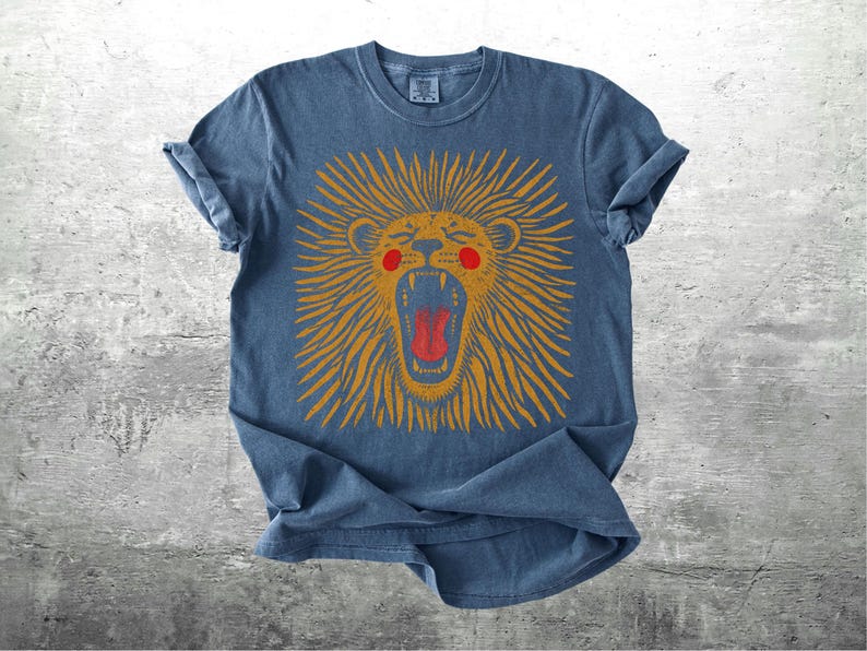 May include: A blue t-shirt featuring a distressed lion design. The lion has an open mouth, orange mane, red tongue, and red cheeks. The shirt has rolled-up sleeves and a crew neck.
