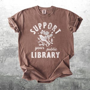 May include: Brown t-shirt with the text "SUPPORT your public LIBRARY" in white. A white illustration of a bird on a branch with flowers is centered above the text. The shirt has rolled-up sleeves and is displayed on a gray surface.
