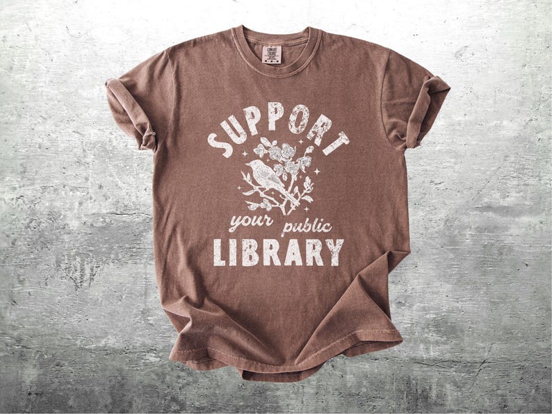 May include: Brown t-shirt with the text "SUPPORT your public LIBRARY" in white. A white illustration of a bird on a branch with flowers is centered above the text. The shirt has rolled-up sleeves and is displayed on a gray surface.