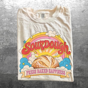May include: A cream-colored t-shirt featuring a graphic of a loaf of sourdough bread centered under a rainbow and clouds. The text reads SOURDOUGH in large red letters and FRESH BAKED HAPPINESS in pink letters on a banner at the bottom.
