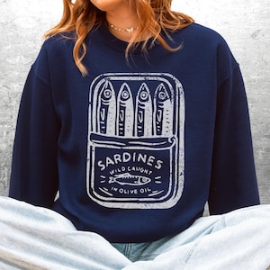 May include: Navy blue sweatshirt with a white graphic of a sardine can. The can features four sardines and the words "Sardines Wild Caught in Olive Oil". The sweatshirt is a casual style.