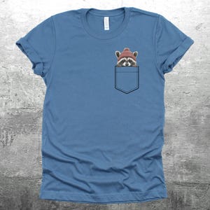 May include: A blue t-shirt with a front pocket featuring a raccoon wearing a red knit hat. The raccoon is peeking out of the pocket.