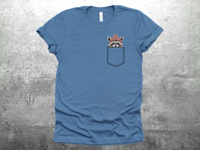 May include: A blue t-shirt with a front pocket featuring a raccoon wearing a red knit hat. The raccoon is peeking out of the pocket.