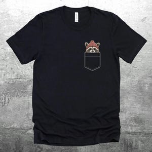 May include: Black t-shirt with a pocket featuring a raccoon wearing a red knit hat. The raccoon is peeking out of the pocket.