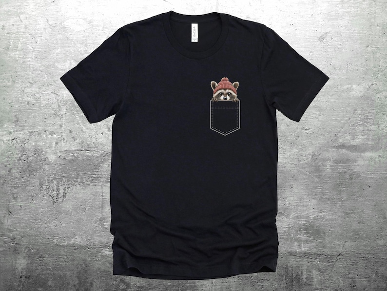 May include: Black t-shirt with a pocket featuring a raccoon wearing a red knit hat. The raccoon is peeking out of the pocket.