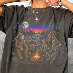 May include: A charcoal gray t-shirt featuring a graphic print of Bigfoot, a moth-like creature playing guitar, an alien, and a swamp creature sitting around a campfire at night. The scene includes a tent, a full moon, and a forest landscape.