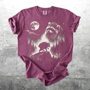 K&ouml;nnte beinhalten: Ein kastanienbraunes Kurzarm-T-Shirt mit einem wei&szlig;en Grafikdruck, der ein Waschb&auml;rengesicht, einen Vollmond und die Silhouette eines Waschb&auml;ren auf einer felsigen Klippe inmitten eines Kiefernwaldes zeigt.