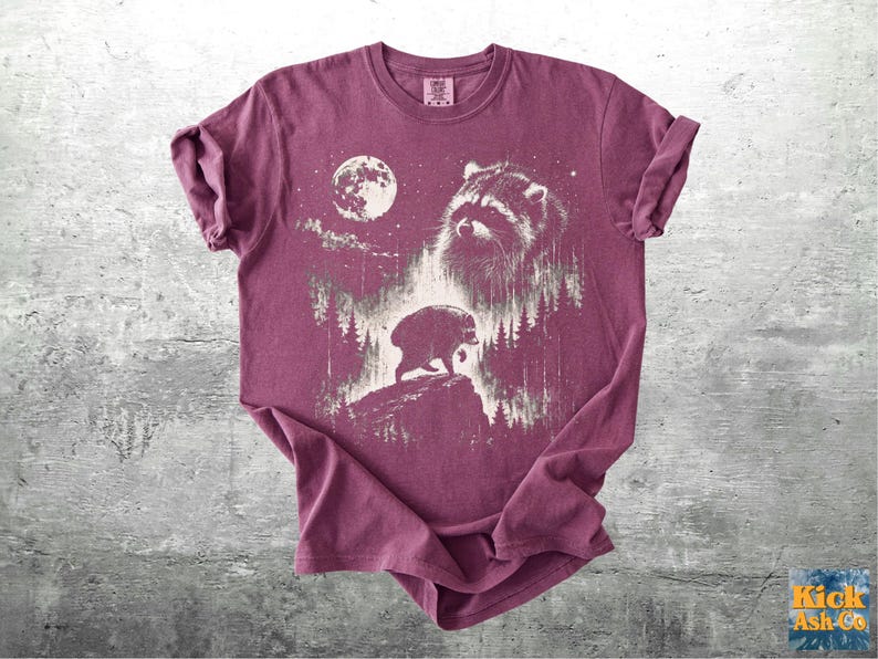 K&ouml;nnte beinhalten: Ein kastanienbraunes Kurzarm-T-Shirt mit einem wei&szlig;en Grafikdruck, der ein Waschb&auml;rengesicht, einen Vollmond und die Silhouette eines Waschb&auml;ren auf einer felsigen Klippe inmitten eines Kiefernwaldes zeigt.