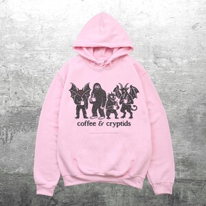 May include: A light pink hoodie with a front pocket and a drawstring hood. The front of the hoodie features a black graphic of several cryptids holding coffee cups, with the words "coffee & cryptids" printed below.