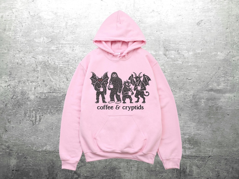 May include: A light pink hoodie with a front pocket and a drawstring hood. The front of the hoodie features a black graphic of several cryptids holding coffee cups, with the words "coffee & cryptids" printed below.
