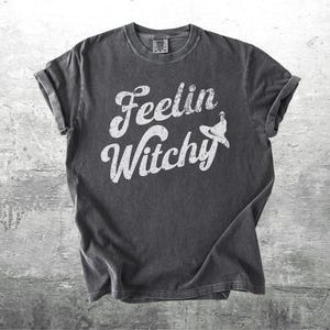 K&ouml;nnte beinhalten: Dunkelgraues T-Shirt mit hochgekrempelten &Auml;rmeln. Das Shirt zeigt die Worte "Feelin Witchy" in einer wei&szlig;en, stilisierten Schriftart, mit einer Hexenhut-Grafik. Das Shirt ist aus weichem Material und f&uuml;r den Freizeitgebrauch geeignet.