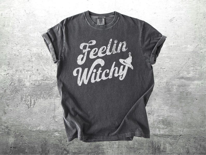 K&ouml;nnte beinhalten: Dunkelgraues T-Shirt mit hochgekrempelten &Auml;rmeln. Das Shirt zeigt die Worte "Feelin Witchy" in einer wei&szlig;en, stilisierten Schriftart, mit einer Hexenhut-Grafik. Das Shirt ist aus weichem Material und f&uuml;r den Freizeitgebrauch geeignet.