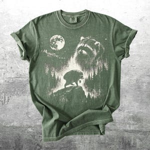 K&ouml;nnte beinhalten: Ein salbeigr&uuml;nes Kurzarm-T-Shirt mit einem wei&szlig;en Grafikdruck, der ein Waschb&auml;rengesicht, einen Vollmond und die Silhouette eines Waschb&auml;ren auf einer felsigen Klippe inmitten eines Kiefernwaldes zeigt. Das Design wirkt strukturiert.