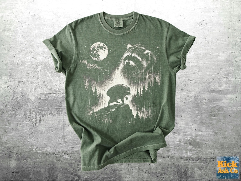 K&ouml;nnte beinhalten: Ein salbeigr&uuml;nes Kurzarm-T-Shirt mit einem wei&szlig;en Grafikdruck, der ein Waschb&auml;rengesicht, einen Vollmond und die Silhouette eines Waschb&auml;ren auf einer felsigen Klippe inmitten eines Kiefernwaldes zeigt. Das Design wirkt strukturiert.