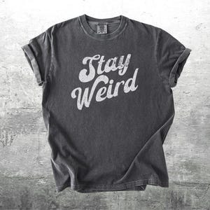 K&ouml;nnte beinhalten: Dunkelgraues T-Shirt mit hochgekrempelten &Auml;rmeln. Das Shirt zeigt den wei&szlig;en Text "Stay Weird" in einer kr&auml;ftigen, stilisierten Schriftart. Das T-Shirt besteht aus einem weichen Material und wird auf einem strukturierten grauen Hintergrund gezeigt.
