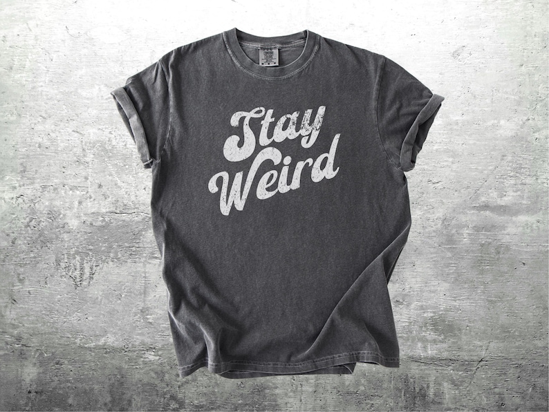 K&ouml;nnte beinhalten: Dunkelgraues T-Shirt mit hochgekrempelten &Auml;rmeln. Das Shirt zeigt den wei&szlig;en Text "Stay Weird" in einer kr&auml;ftigen, stilisierten Schriftart. Das T-Shirt besteht aus einem weichen Material und wird auf einem strukturierten grauen Hintergrund gezeigt.