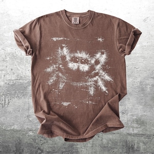 Op de afbeelding: Bruin t-shirt met een witte spinnenafbeelding. Het t-shirt heeft een ronde hals en opgerolde korte mouwen. De stof heeft een vervaagde uitstraling. Het spinnenontwerp is groot en gedetailleerd. Het t-shirt ligt plat op een getextureerd, grijs oppervlak.