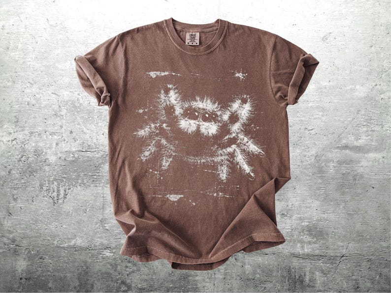 Op de afbeelding: Bruin t-shirt met een witte spinnenafbeelding. Het t-shirt heeft een ronde hals en opgerolde korte mouwen. De stof heeft een vervaagde uitstraling. Het spinnenontwerp is groot en gedetailleerd. Het t-shirt ligt plat op een getextureerd, grijs oppervlak.